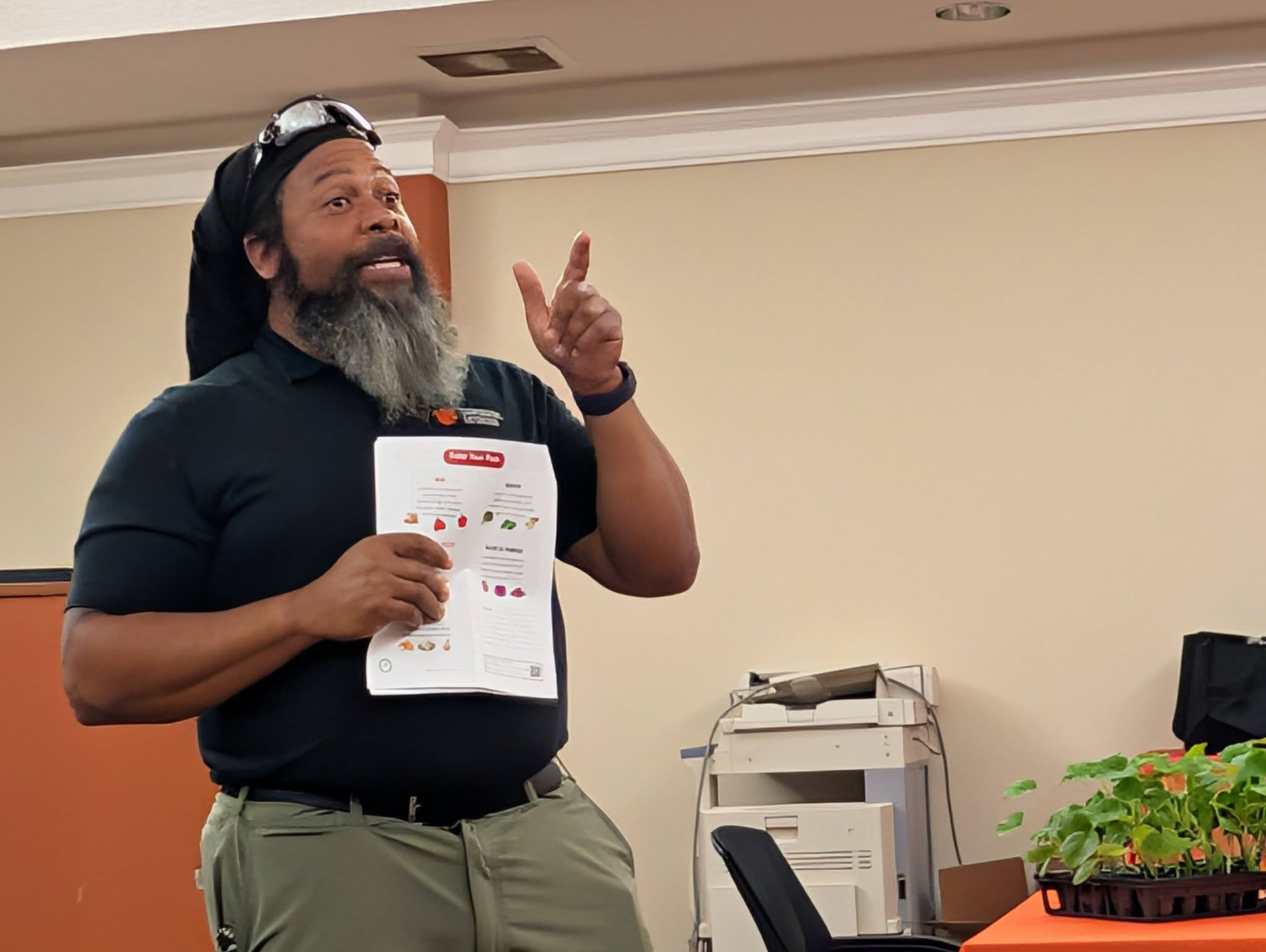 A man stands indoors holding a printed sheet, gesturing with his other hand, wearing a black shirt and cap, with a copier and table visible behind him.