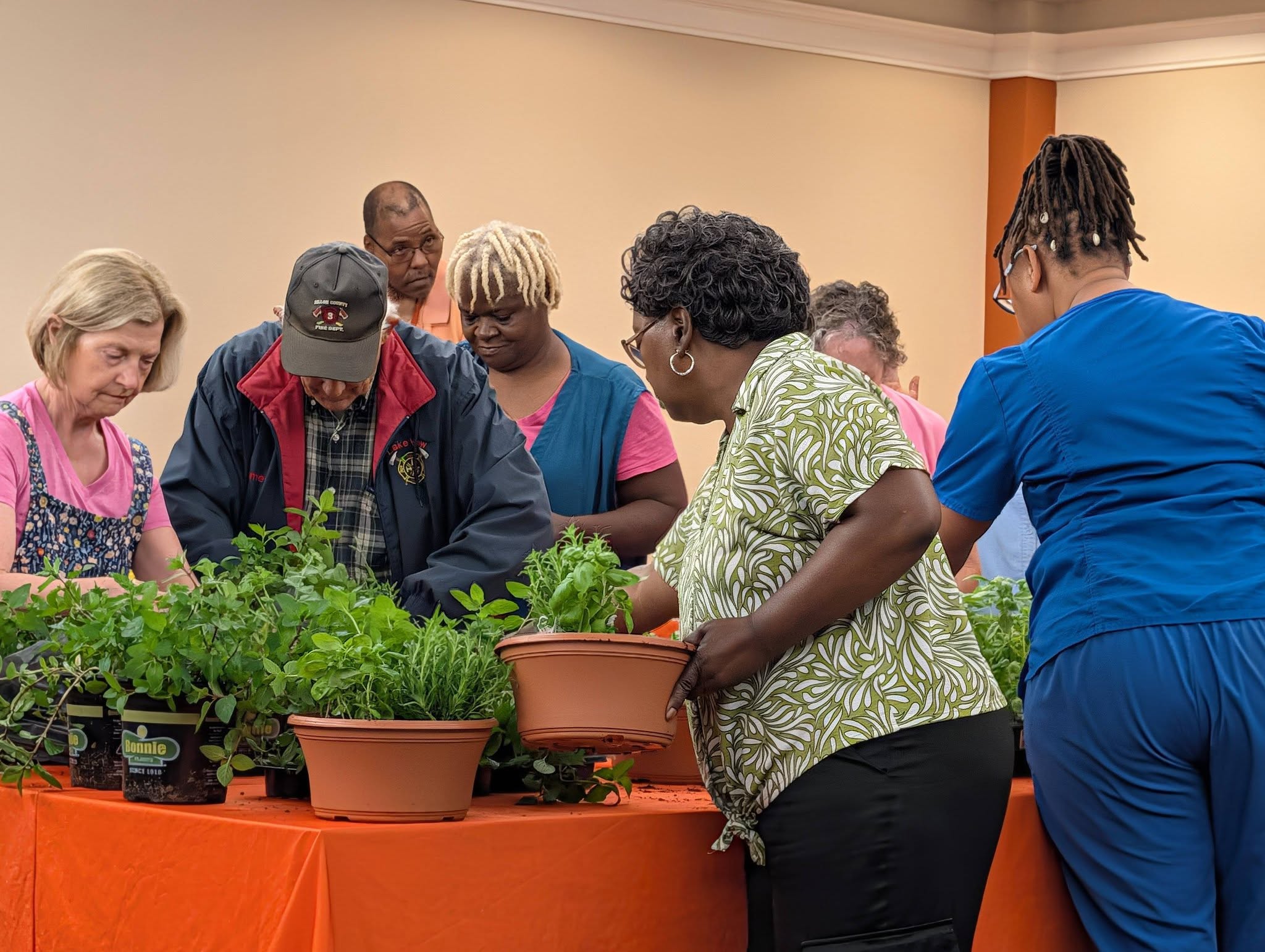 A group of people gathers around a table with potted plants, engaging in a hands-on gardening activity indoors.
