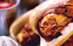Close-up of grilled sausages in buns with sauerkraut and ketchup, served on a plate. Blurred background shows bowls of mustard and ketchup with wooden spoons.