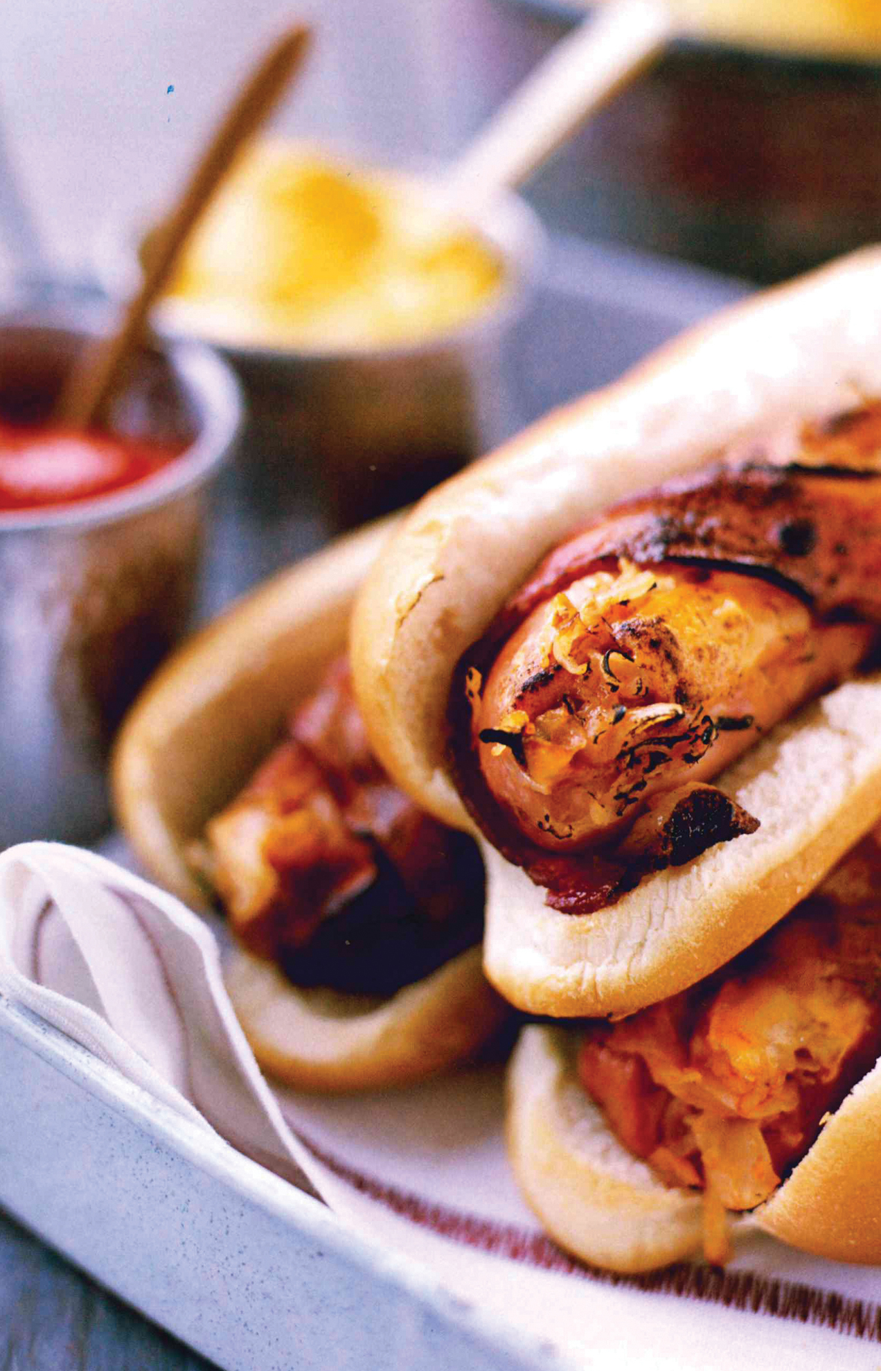 Close-up of grilled sausages in buns with sauerkraut and ketchup, served on a plate. Blurred background shows bowls of mustard and ketchup with wooden spoons.
