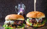 Two cheeseburgers with leafy greens and sauce on a wooden board, accompanied by a small glass bottle of sauce, rustic background.