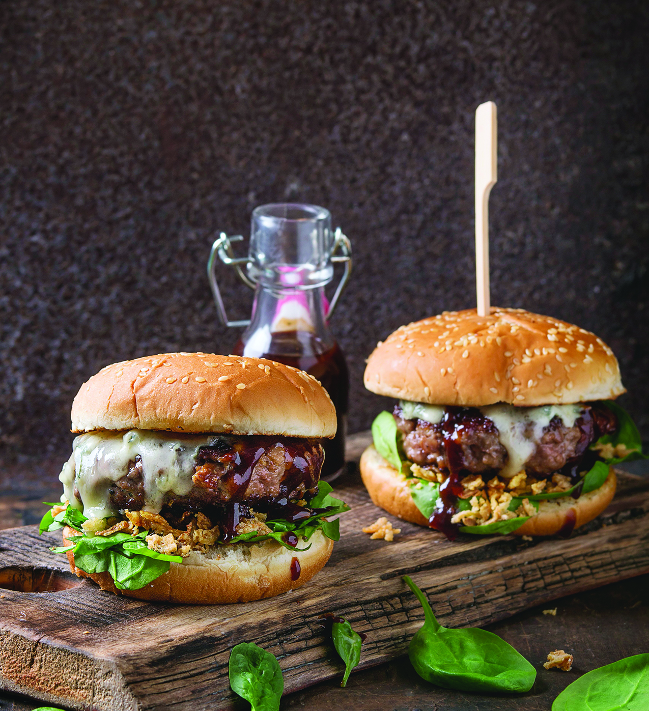 Two cheeseburgers with leafy greens and sauce on a wooden board, accompanied by a small glass bottle of sauce, rustic background.