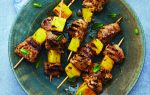 Grilled meat and pineapple skewers on a green plate, garnished with mint leaves, served with a small bowl of dipping sauce containing sliced red chili.