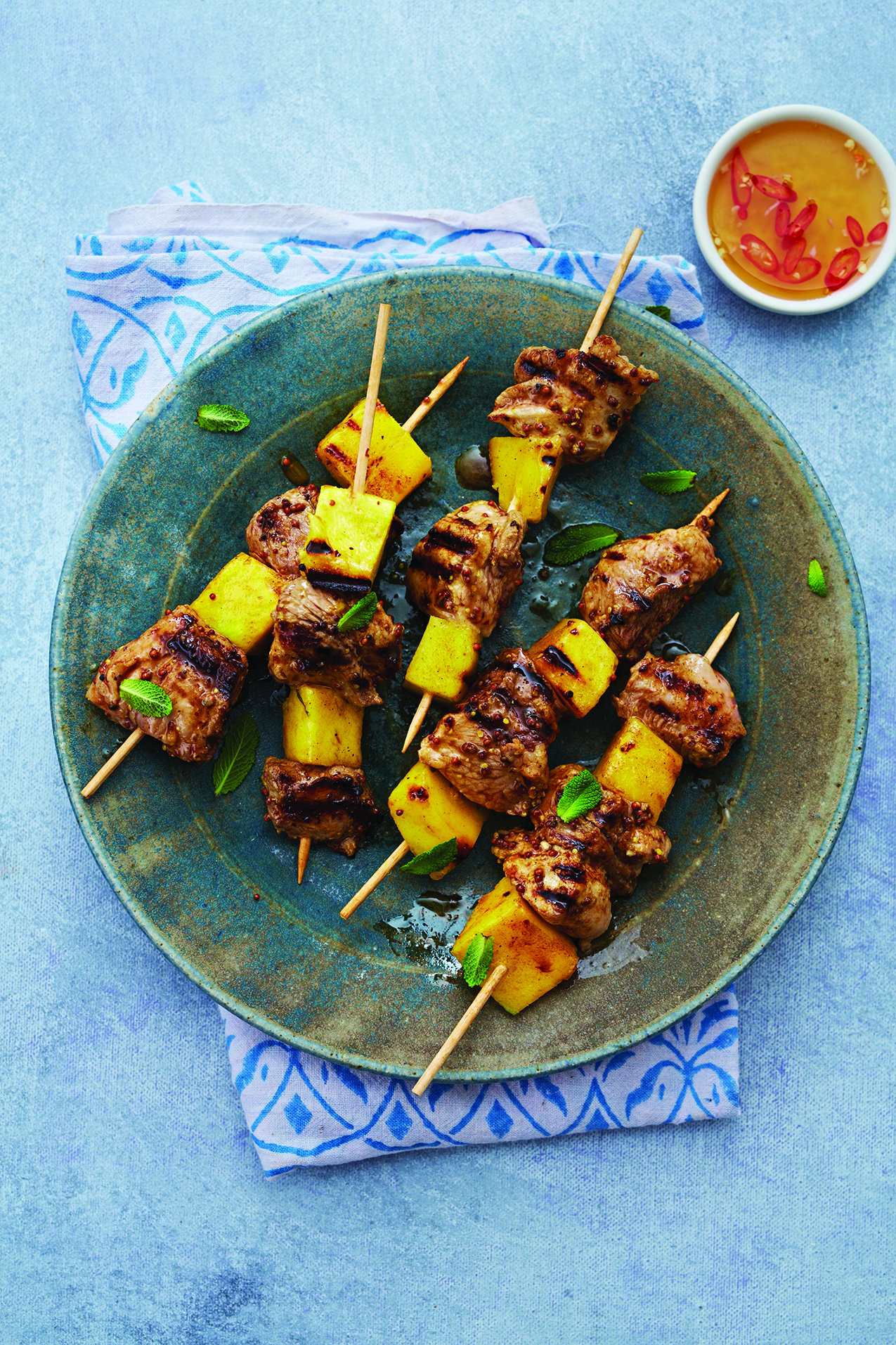 Grilled meat and pineapple skewers on a green plate, garnished with mint leaves, served with a small bowl of dipping sauce containing sliced red chili.