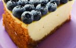 A slice of cheesecake topped with fresh blueberries and powdered sugar, served on a purple plate with a floral background.