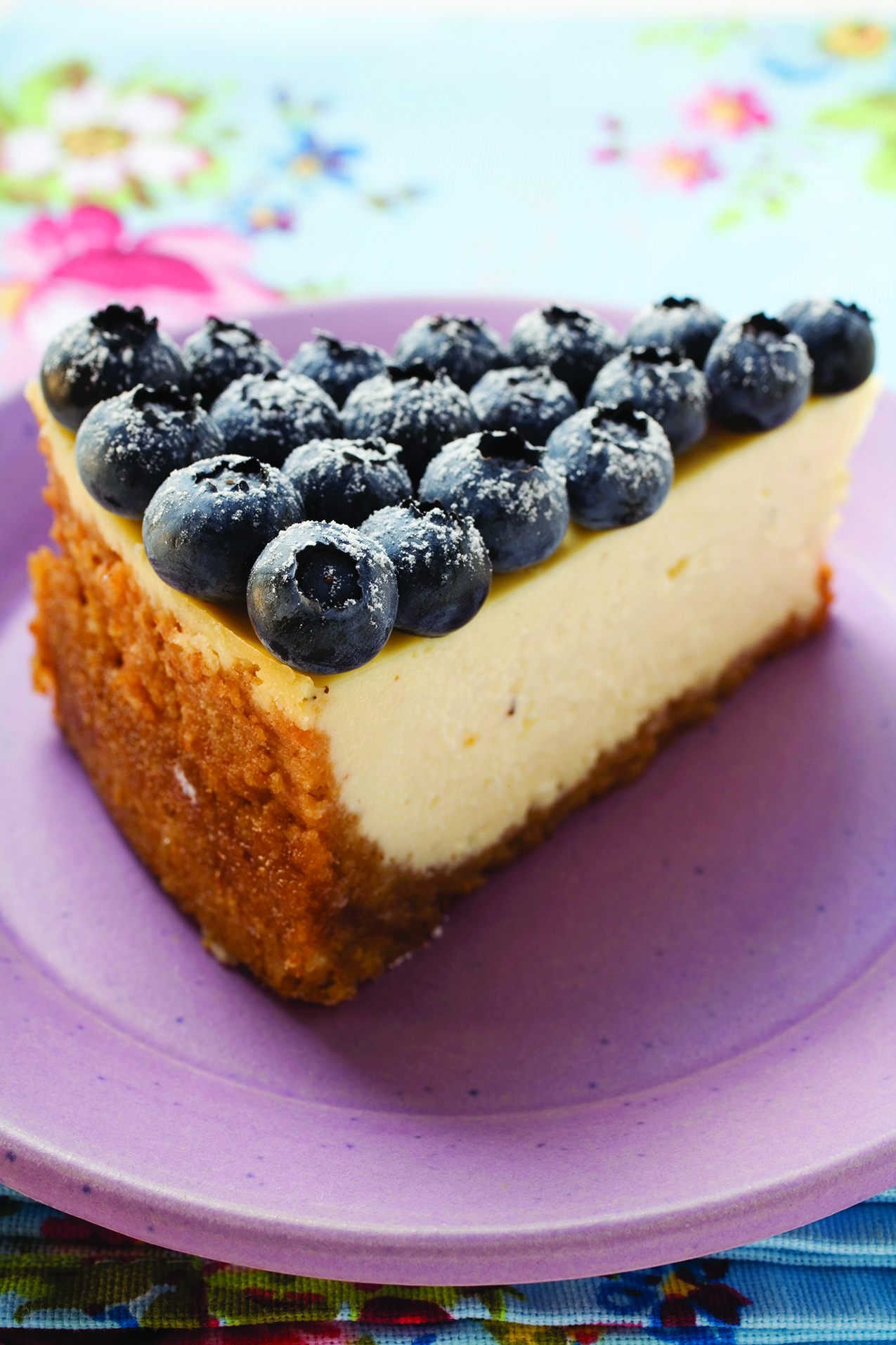 A slice of cheesecake topped with fresh blueberries and powdered sugar, served on a purple plate with a floral background.