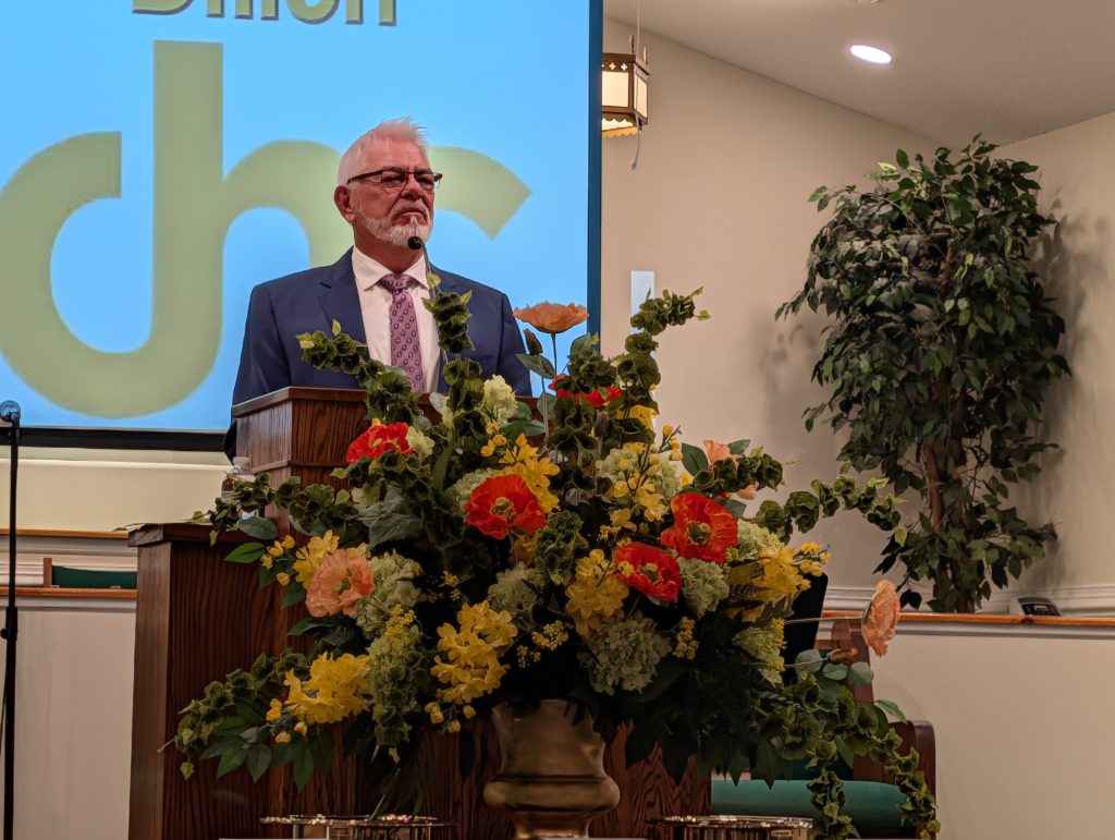 older man in a suit stands at a church podium with a large flower arrangement, giving a speech or sermon.