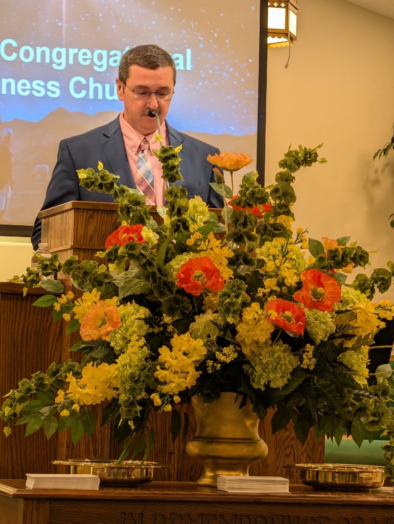 A man in a suit stands at a podium with a large floral arrangement in front of him, in a church with a projection screen behind.