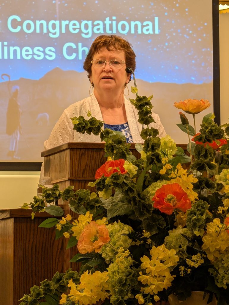 A woman speaks at a church podium with a microphone headset, standing behind colorful flowers, with a projected church message in the background.
