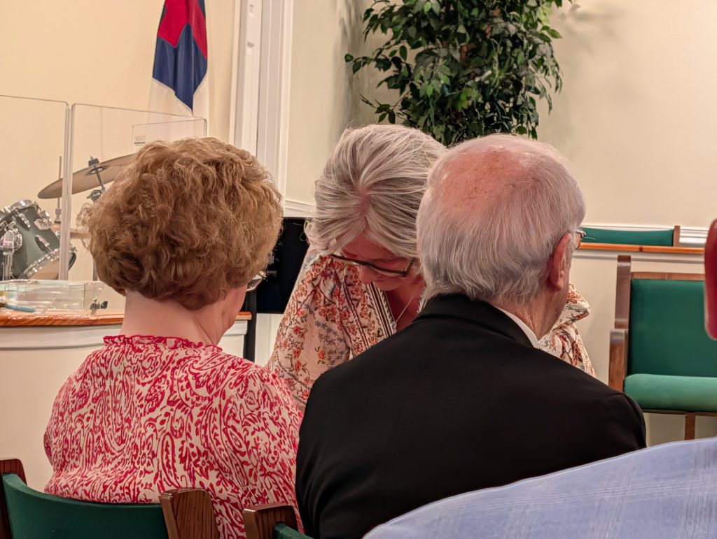 Three older adults sit together in a church, talking closely. A drum set, flag, and green chairs are visible in the background.