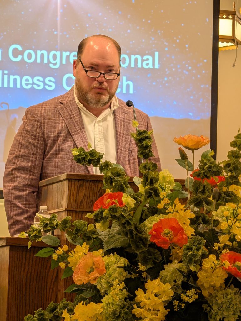 Man wearing glasses stands at a podium adorned with colorful flowers, speaking in front of a projected church presentation.