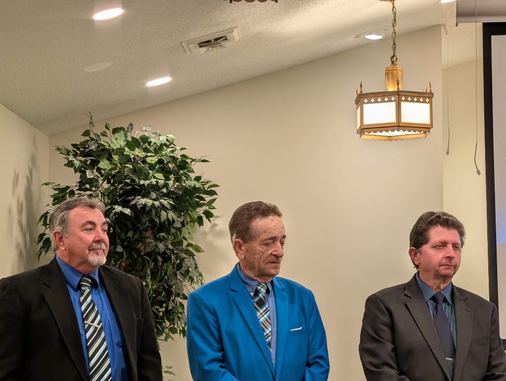Three men in suits stand indoors near a potted plant and a hanging lamp, with neutral expressions, in a formal setting.