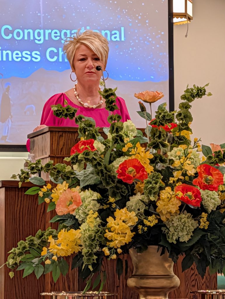 Woman speaking at a podium with a microphone, large floral arrangement in front, and a presentation screen with text in the background.