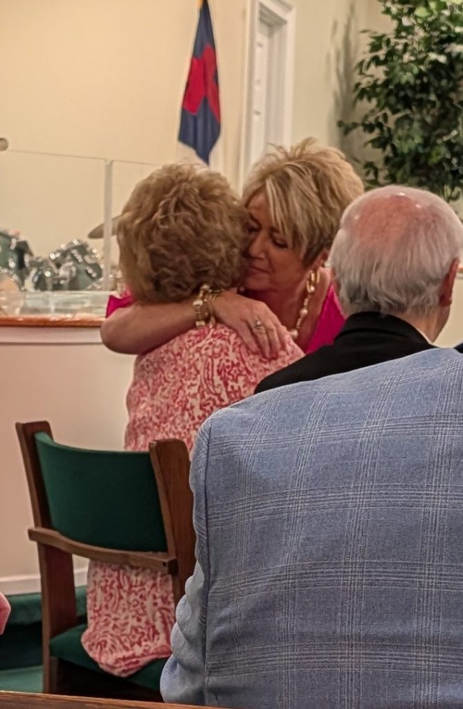 Two women hugging in a church pew, one wearing pink. An elderly man sits in the foreground. A flag and plants are in the background.
