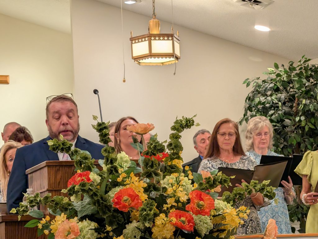 A group of people singing in a church, standing behind a podium decorated with colorful flowers, under a hanging light fixture.