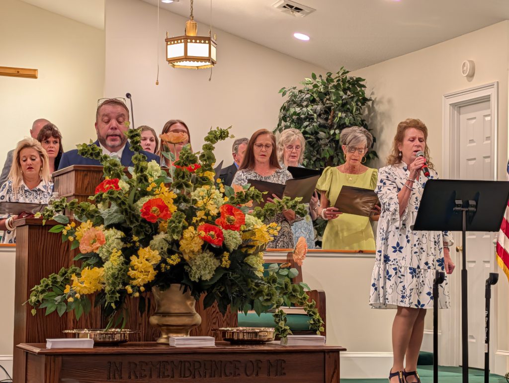 A church choir sings during a service, with a woman singing solo at a microphone. Large flower arrangement and communion trays are in the foreground.