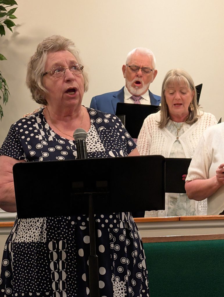 elderly woman sings into a microphone at a church service, accompanied by a small choir in the background.
