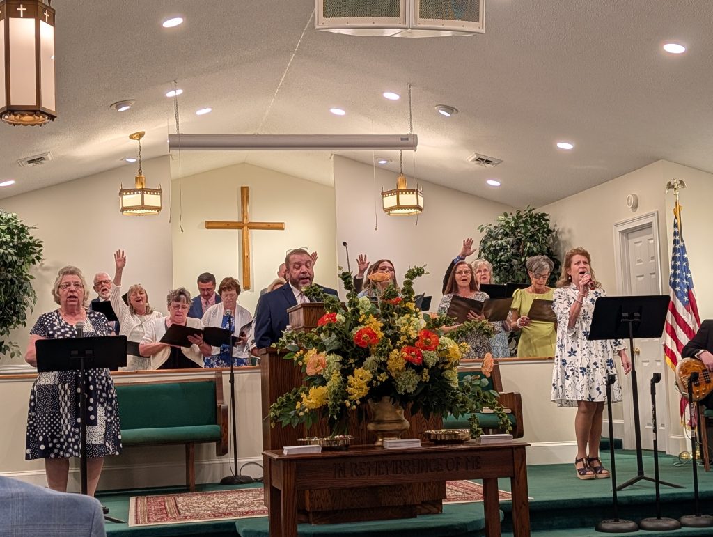 A choir sings in a church with floral arrangements, a wooden cross, and American flag, while several members hold songbooks and a woman sings into a microphone.