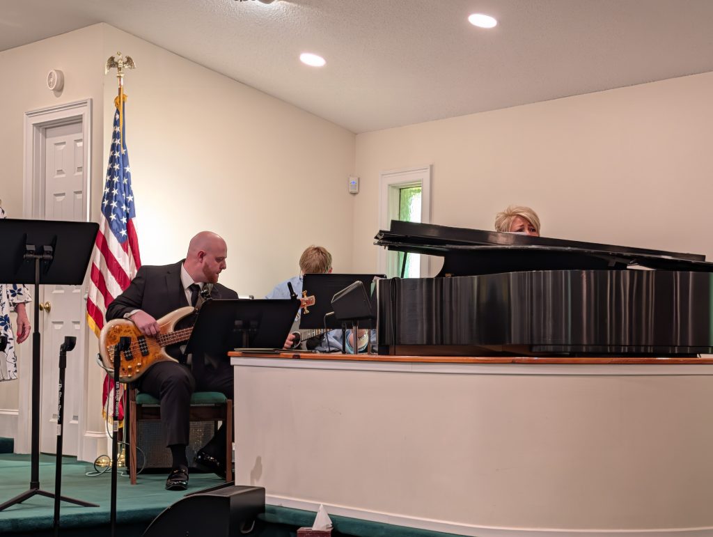 Musicians perform inside a church, one playing a guitar and another at a grand piano, with the American flag in the background.