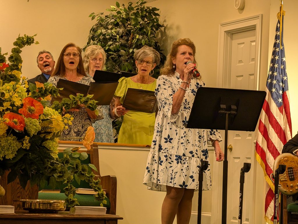 A group of adults sing in a church, one woman leads with a microphone. Floral arrangements and an American flag are visible in the background.