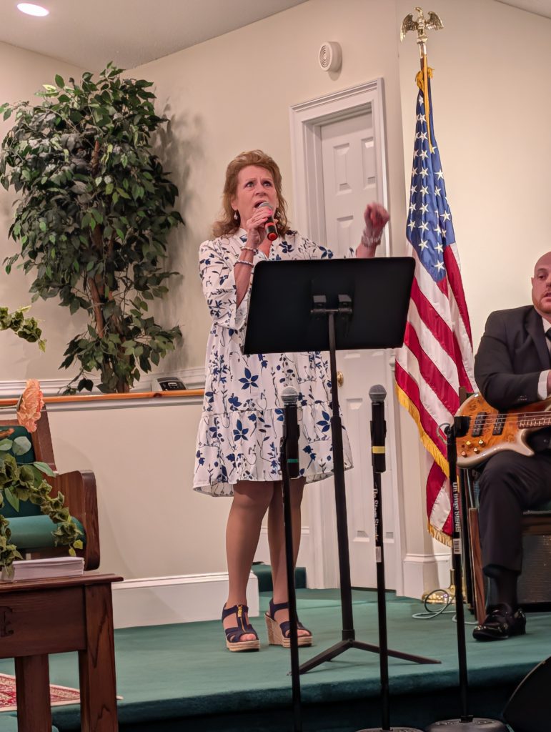 A woman in a floral dress sings into a microphone on a stage with an American flag and musician playing guitar in the background.