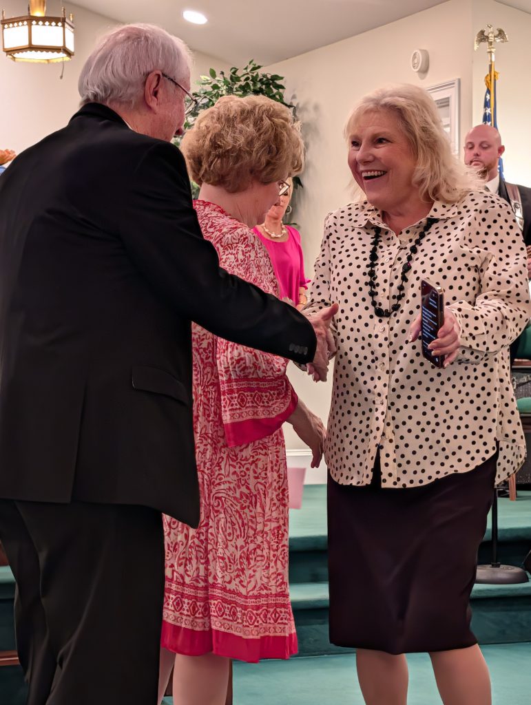 Two women and a man greet and smile inside a well-lit room, possibly at a formal event or gathering, with others present in the background.