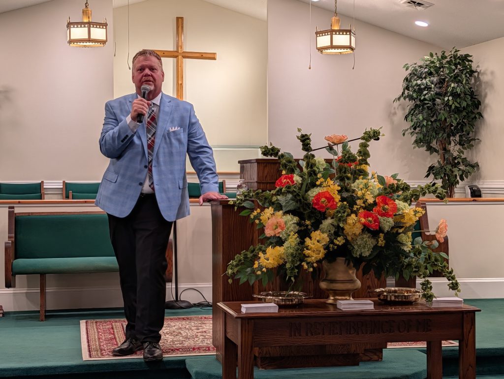 A man in a blue jacket speaks into a microphone in a church, with floral arrangements and a wooden table inscribed “In Remembrance of Me.”