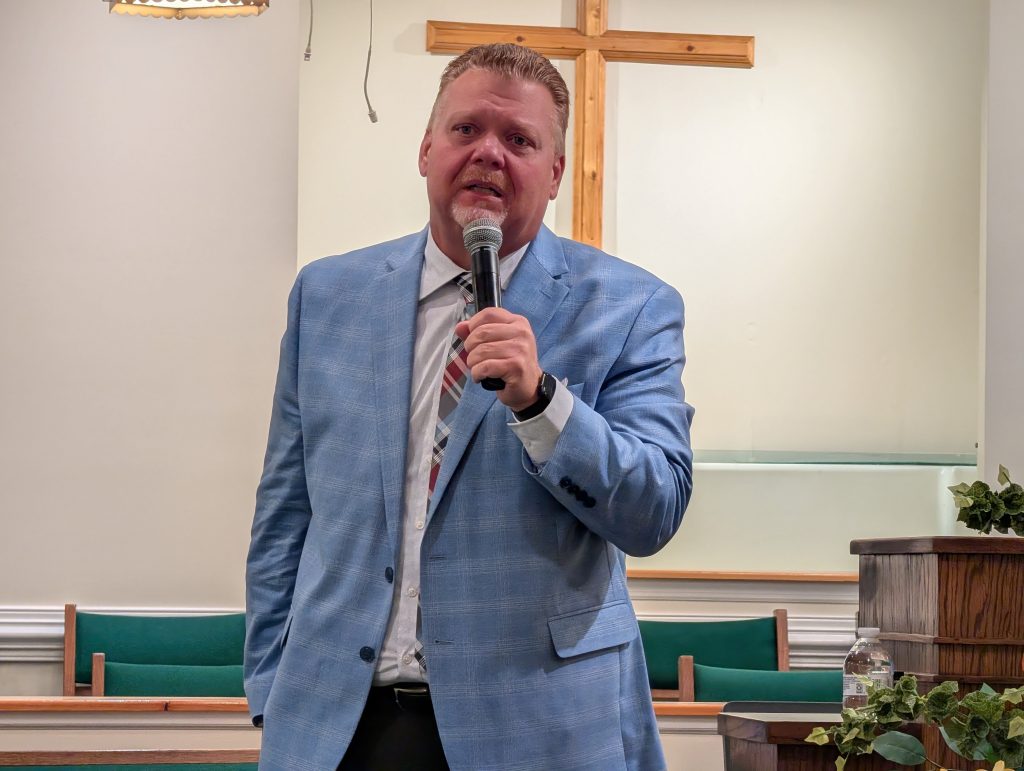 Man in a blue suit speaking into a microphone at a church, standing in front of a wooden cross and church pews.