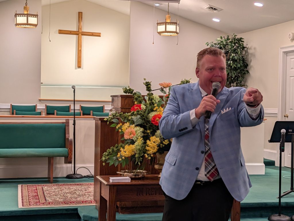 Man in a blue suit speaking and pointing with a microphone inside a church, with a wooden cross and floral arrangement in the background.