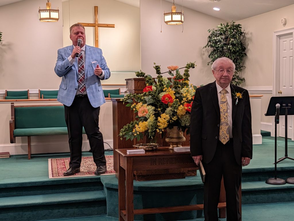Two men in suits stand at the front of a church; one speaks into a microphone, the other stands by a pulpit with flowers and a Bible.