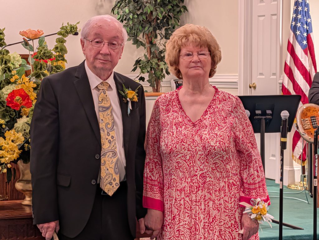 Older couple standing together indoors, the man in a suit and the woman in a patterned dress. Floral decorations and an American flag are visible in the background.