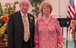 Older couple standing together indoors, the man in a suit and the woman in a patterned dress. Floral decorations and an American flag are visible in the background.