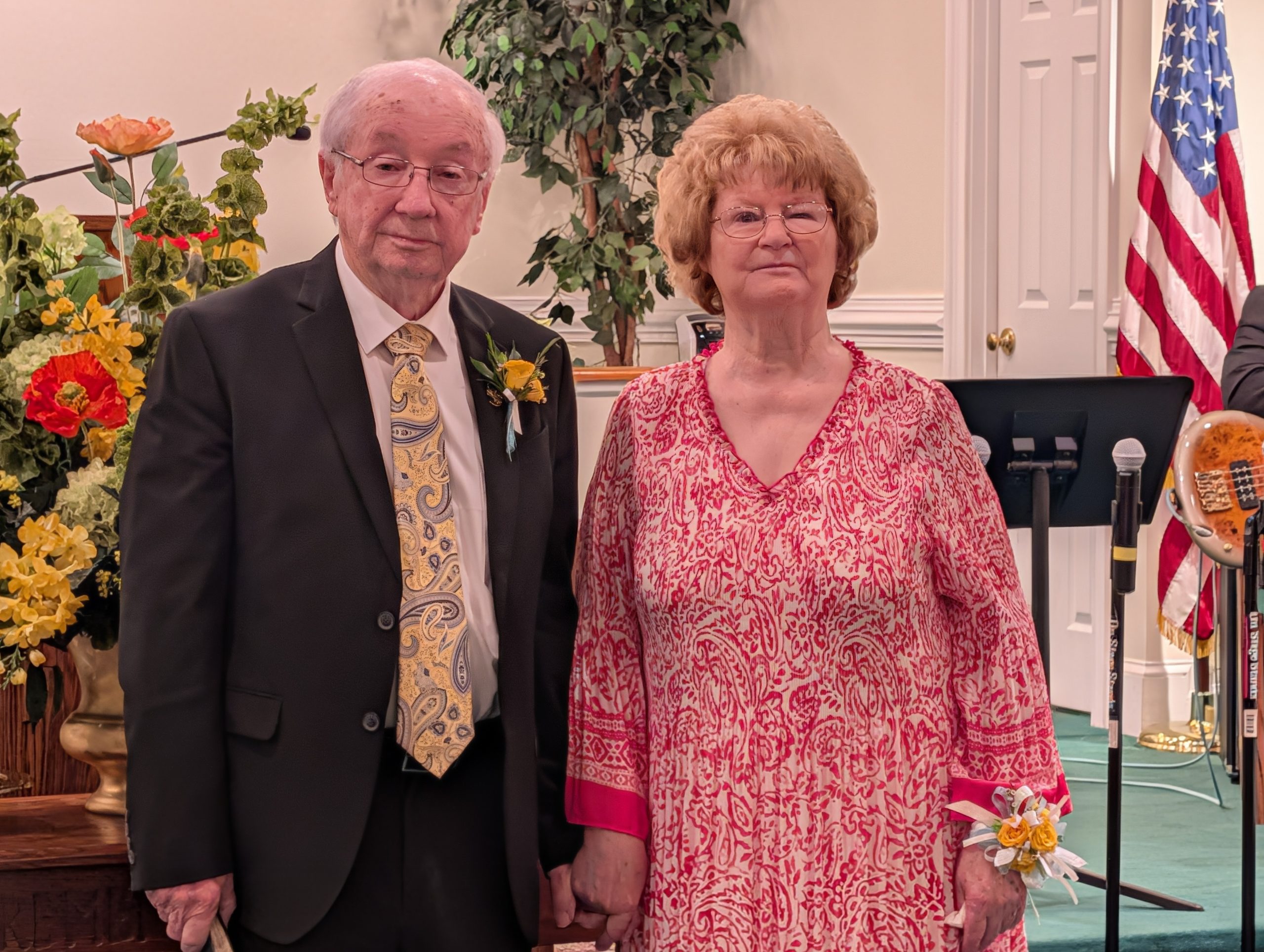 Older couple standing together indoors, the man in a suit and the woman in a patterned dress. Floral decorations and an American flag are visible in the background.