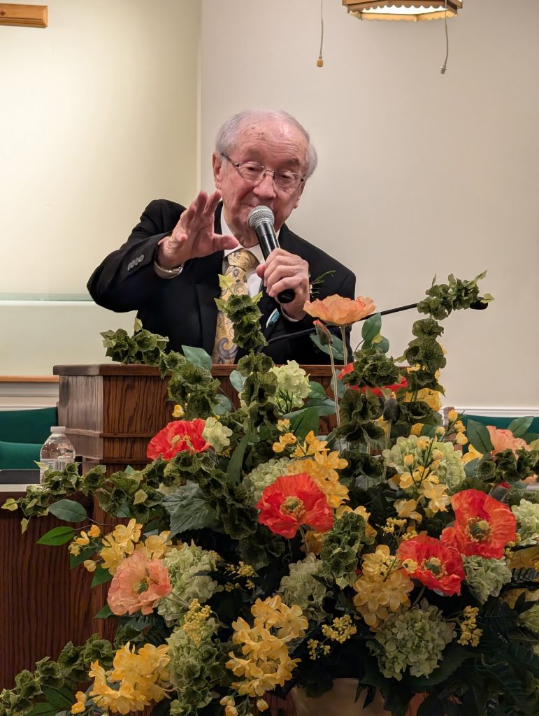 elderly man in a suit speaks into a microphone behind a podium, with a large, colorful floral arrangement in the foreground.