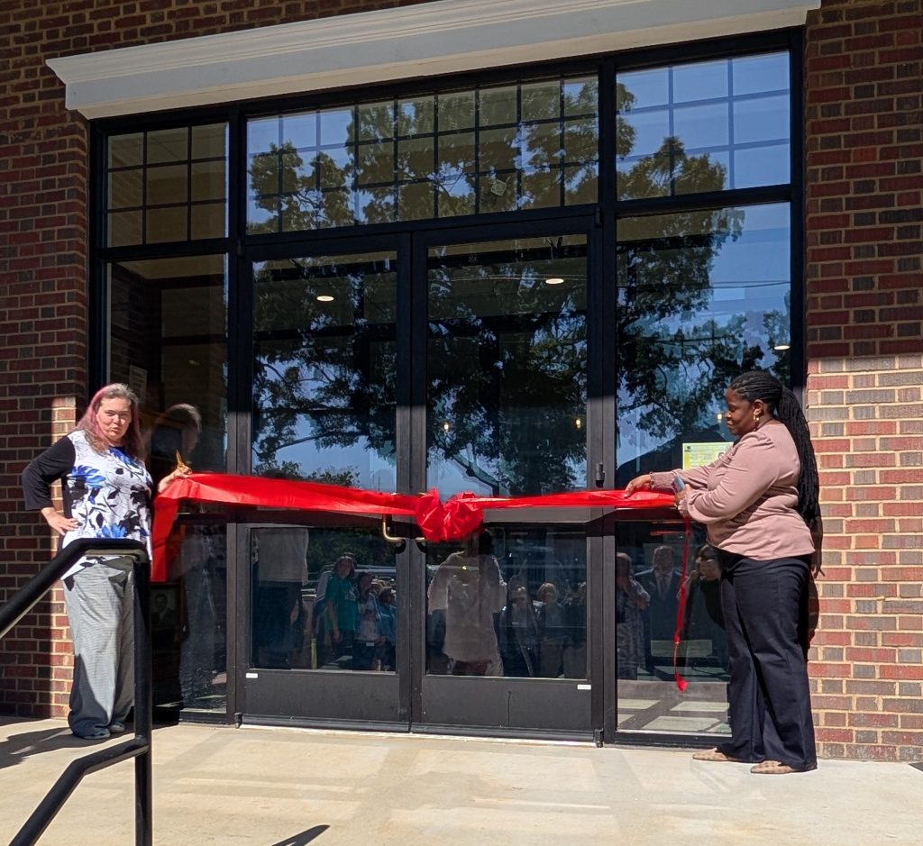 Two women cut a large red ribbon in front of glass doors during a ribbon-cutting ceremony, with people visible inside the building.