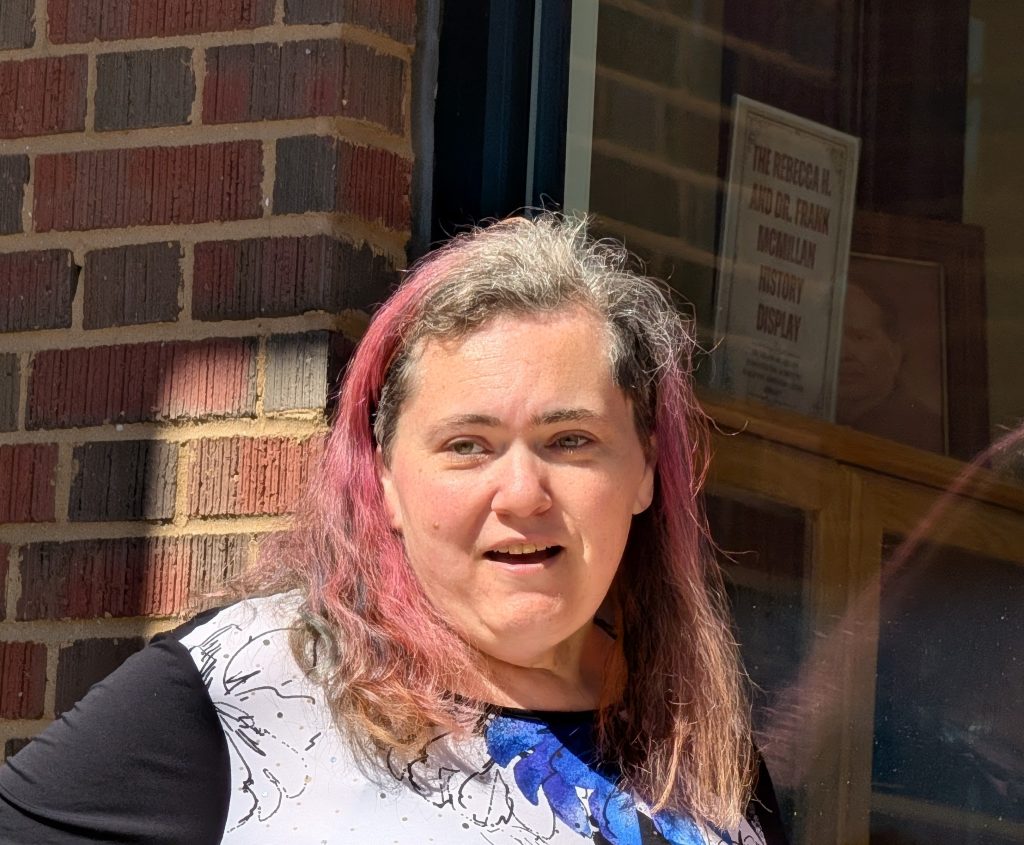 A woman with shoulder-length, wavy hair, partly dyed pink, stands outside near a brick wall, sunlit, with a window reflecting a historical display sign behind her.