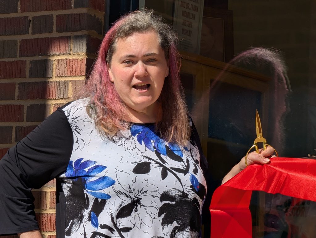 Woman with pink hair holds gold scissors, preparing to cut a red ribbon at an event, standing next to a brick wall and window.