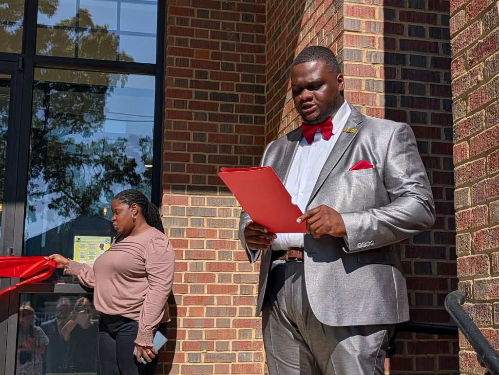 A man in a gray suit reads from a red folder, while a woman holds a red ribbon outside a brick building.