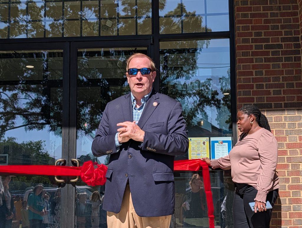 A man in sunglasses stands speaking at a ribbon-cutting event outside a brick building, with a woman holding the red ribbon nearby.
