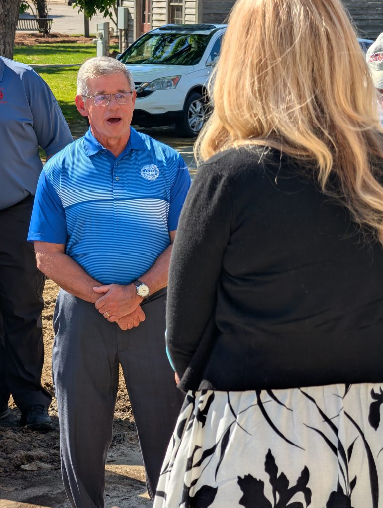 older man wearing glasses and a blue polo shirt speaks outdoors to a woman with long blond hair, while others stand nearby.