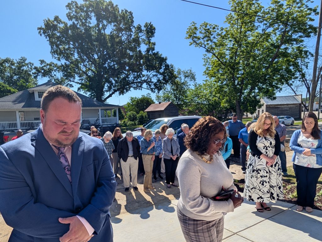 A group of people stand outside in daylight with heads bowed, appearing to pray or observe a moment of silence, surrounded by trees and houses.