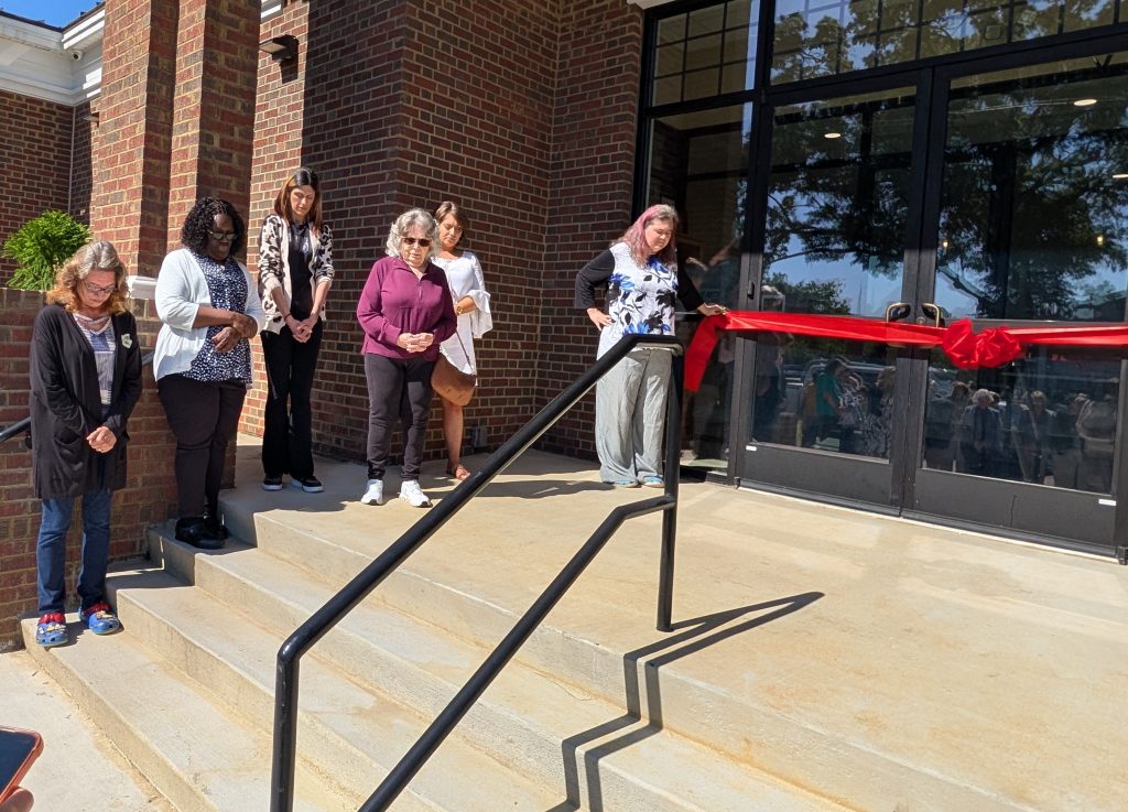 A group of women stand on steps with heads bowed, next to a large red ribbon in front of a building's glass doors, during a ribbon-cutting ceremony.