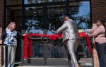 Three people participate in a ribbon-cutting ceremony outside a building, one man in a suit is cutting a large red ribbon, while two women hold each end.