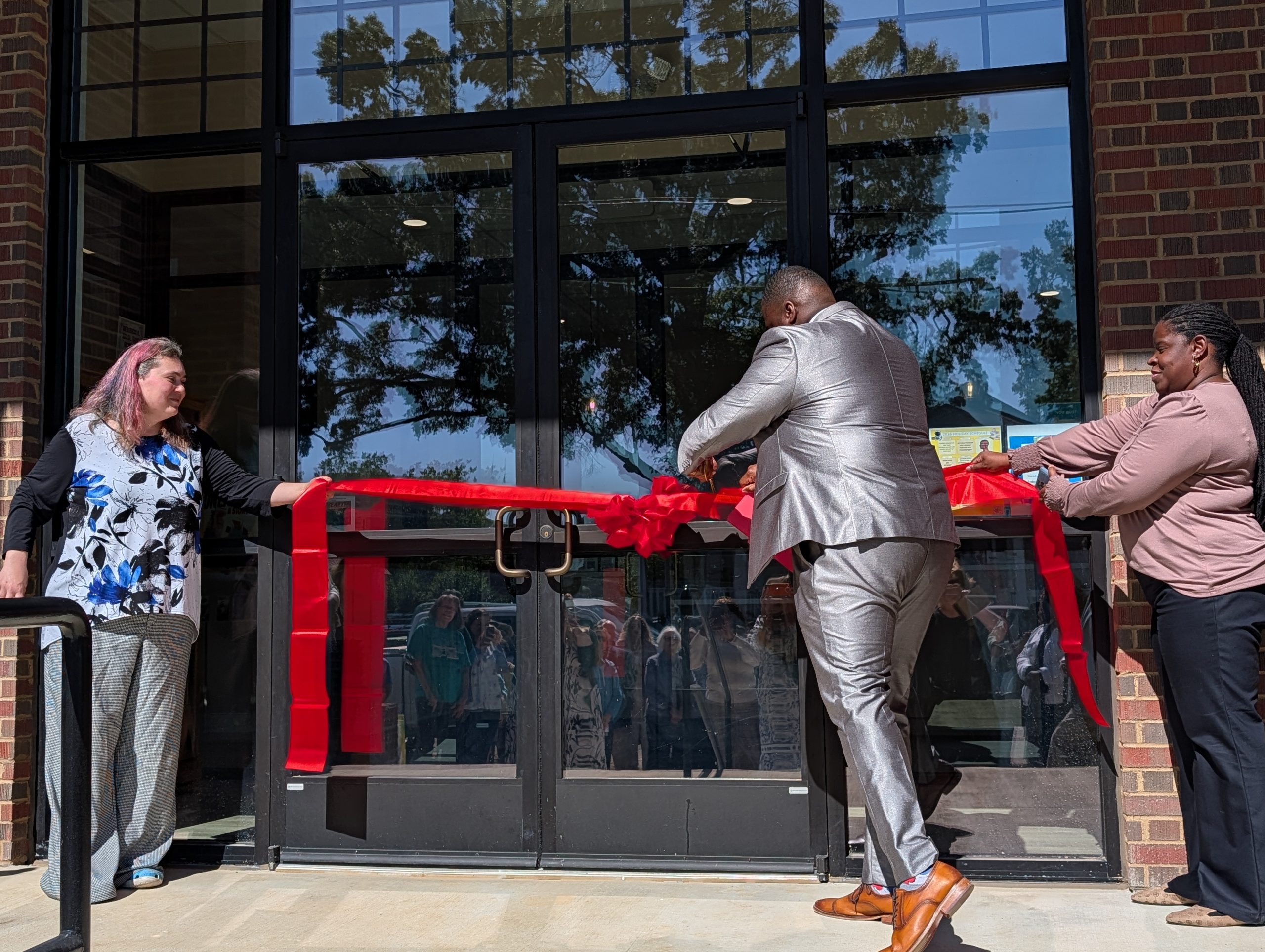 Three people participate in a ribbon-cutting ceremony outside a building, one man in a suit is cutting a large red ribbon, while two women hold each end.