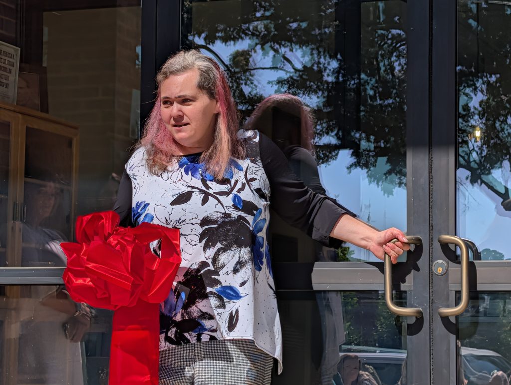 Person with pink-streaked hair holds a door handle and large red ribbon, standing in sunlight near glass doors, possibly at a ribbon-cutting event.