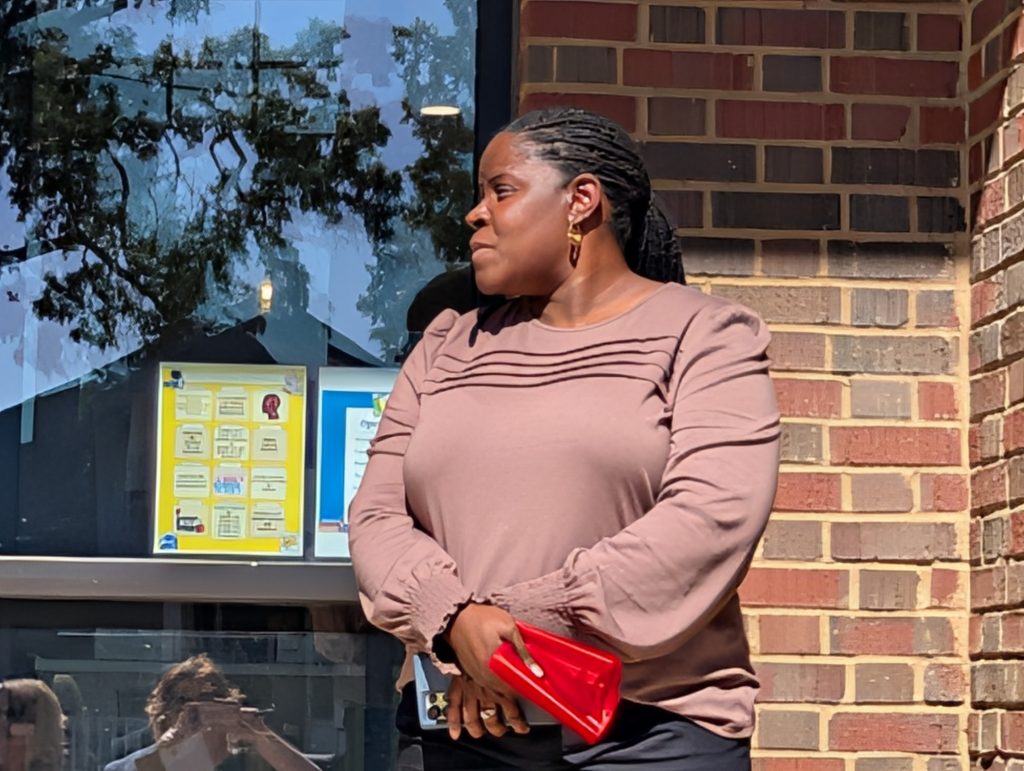 A woman in a mauve blouse stands outside a brick building, holding a red wallet, with sunlight and reflections visible in the window behind her.