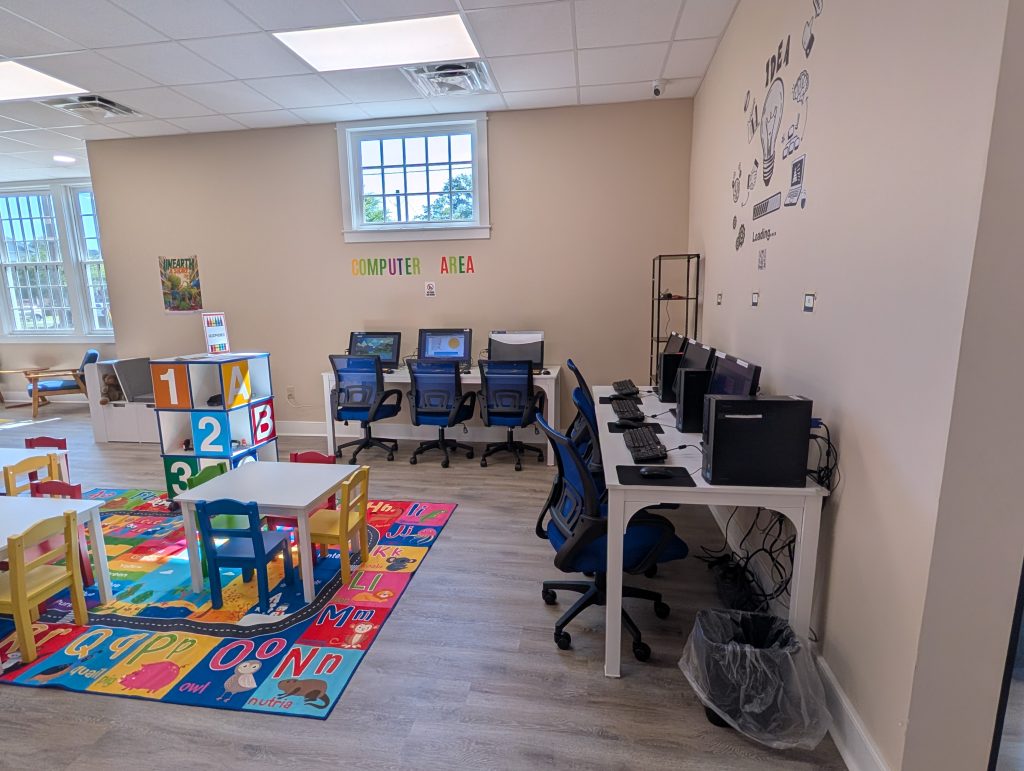 Children’s computer area in a classroom with colorful chairs, tables, alphabet rug, computers on desks, and educational decorations on the wall.
