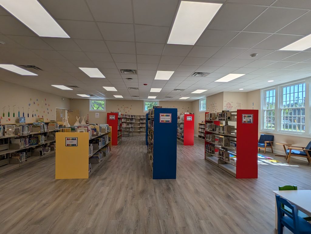 Children’s library with colorful bookshelves, bright lighting, wooden floors, large windows, and seating areas. Books and decorations create a welcoming reading environment for kids.