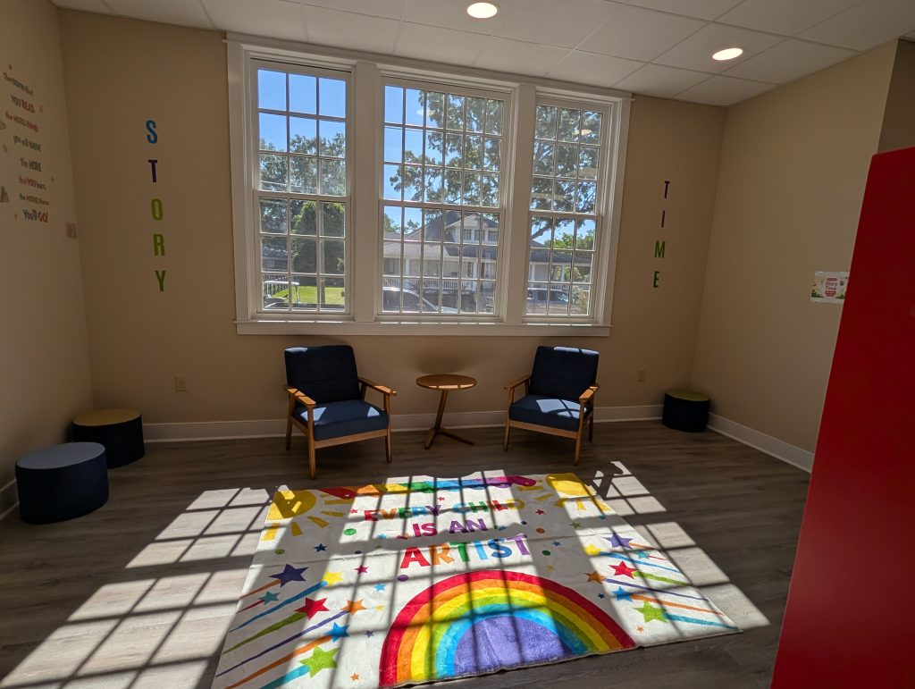 Bright reading room with large windows, two chairs, small tables, and a colorful rug featuring a rainbow and stars. "Story Time" letters are on the walls.