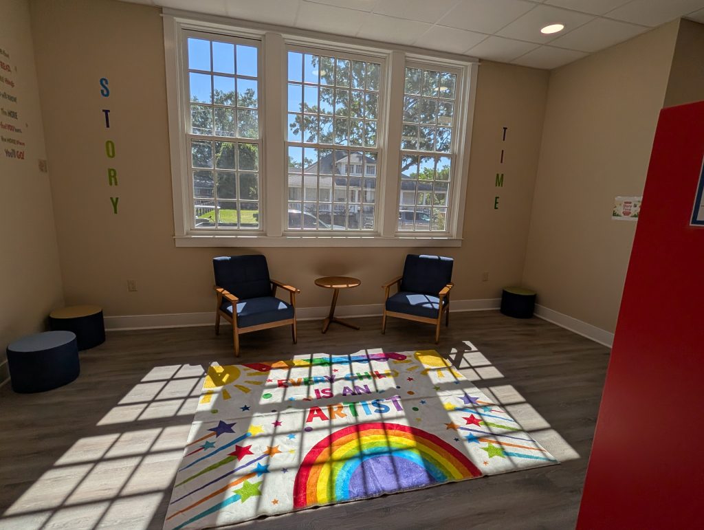 Bright reading nook with two chairs, colorful rug featuring a rainbow and “Artist” text, large windows, and “Story Time” letters on the wall.
